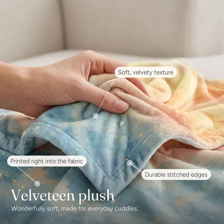 Close-up of a hand holding the velveteen plush Everyday Love Blanket, showing the soft short pile, the printed design and the stitched edge